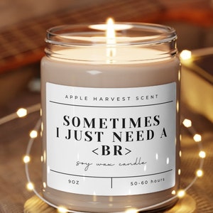 May include: A soy wax candle with a brown glass jar and a white label that reads "Sometimes I just need a <br> soy wax candle" and "Apple Harvest Scent". The candle is sitting on a wooden coaster and is surrounded by white string lights.