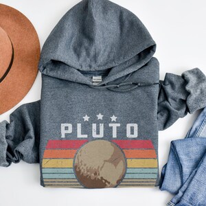 May include: A heather gray Pluto hoodie with a retro design. The design features the word "PLUTO" above a graphic of the planet with a colorful striped background. A brown hat and blue jeans are also in the image.
