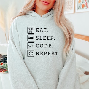 May include: A light gray hoodie with the words "EAT. SLEEP. CODE. REPEAT." in black block letters. The text is arranged in a vertical list with corresponding icons: a fork and knife, a bed, code brackets, and a circular arrow.