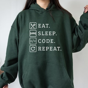 May include: Dark green hoodie with white text and icons. The text reads "EAT. SLEEP. CODE. REPEAT." with corresponding icons for each word: a fork and knife, a bed, code brackets, and a circular arrow.