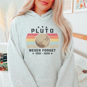 May include: A light gray hoodie with a retro design featuring the word "PLUTO" above a graphic of the planet. Below the planet graphic, the text reads "NEVER FORGET 1930 - 2006". The design incorporates a sunset color scheme.