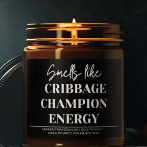 May include: A lit candle in a glass jar with a black label. The label reads "Smells like Cribbage Champion Energy." The jar is brown with a silver lid. The candle is hand-poured with vegan soy wax.