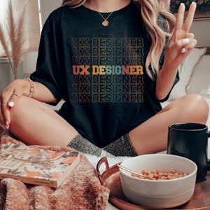 May include: A black t-shirt with the text "UX Designer" repeated in a rainbow gradient pattern. The shirt is worn by a person sitting on a bed with a bowl of cereal and a mug.