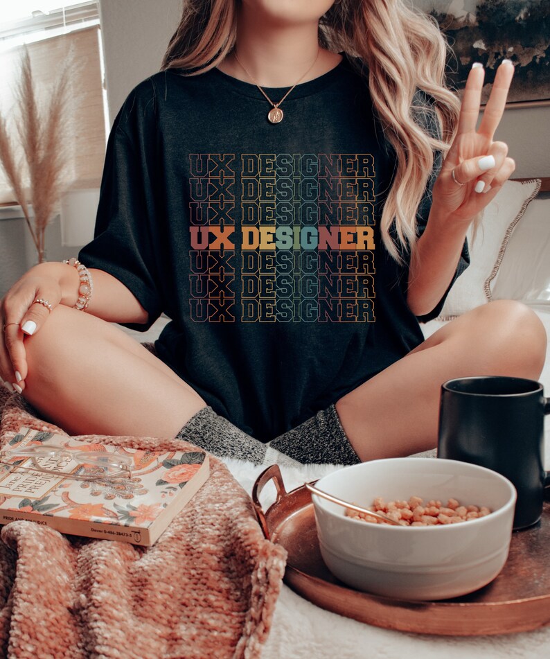 May include: A black t-shirt with the text "UX Designer" repeated in a rainbow gradient pattern. The shirt is worn by a person sitting on a bed with a bowl of cereal and a mug.