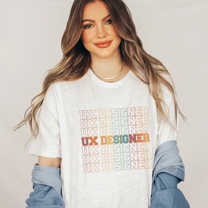 May include: A white t-shirt with the text "UX Designer" repeated in a rainbow color pattern.