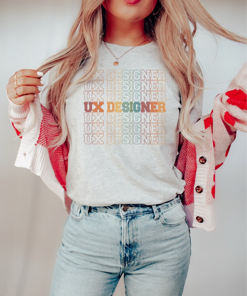 May include: A woman wearing a grey t-shirt with the text "UX Designer" repeated in a rainbow pattern. She is also wearing blue jeans and a white cardigan with red accents.