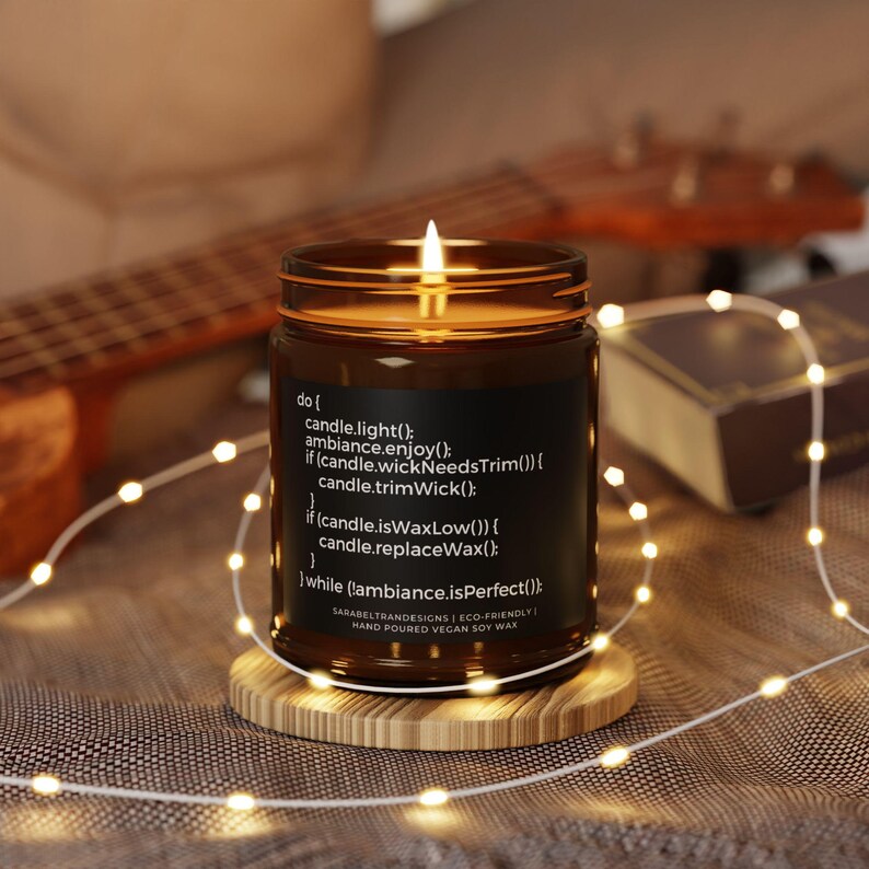May include: A brown glass candle jar with a black label that reads "do { candle.light(); ambiance.enjoy(); if (candle.wickNeedsTrim()) { candle.trimWick(); if (candle.isWaxLow()) { candle.replaceWax(); } while (!ambiance.isPerfect()); } SARABELTRANDESIGNS | ECO-FRIENDLY | HAND POURED VEGAN SOY WAX". The candle is lit and the flame is visible.