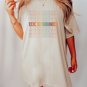 May include: A beige oversized t-shirt with the text "UX Designer" repeated in a rainbow pattern.