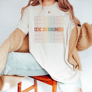 May include: A white t-shirt with the text "UX Designer" repeated in a rainbow gradient pattern.