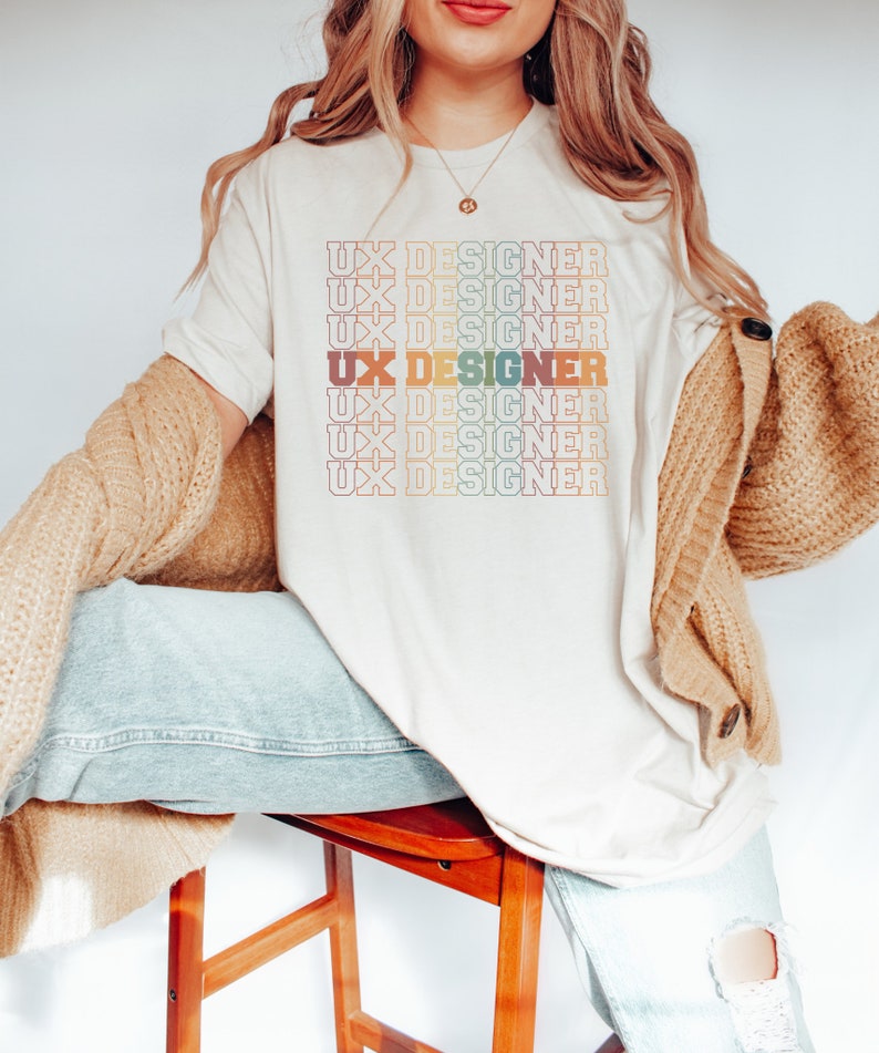 May include: A white t-shirt with the text "UX Designer" repeated in a rainbow gradient pattern.