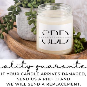 May include: A white candle in a glass jar with a white label that says "CDD SARABELTRANDESIGNS UDD". The candle is sitting on a wooden tray with green plants.