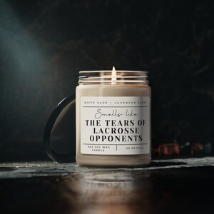Puede incluir: Una vela de cera de soja beige con una etiqueta blanca que dice "Smells like THE TEARS OF LACROSSE OPPONENTS" con los detalles "WHITE SAGE LAVENDER SCENT", "9OZ SOY WAX CANDLE" y "50-60 hours".