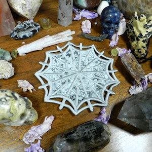 May include: A circular, silver-colored embroidered patch featuring a detailed spider web design with radiating lines and interconnected segments, showcasing a metallic sheen and precise stitching throughout the entire web pattern.