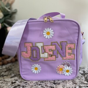 May include: A lavender lunch bag with a zipper closure and a personalized name "JOLENE" in pink, white, and gold letters. The bag is decorated with embroidered white daisies.