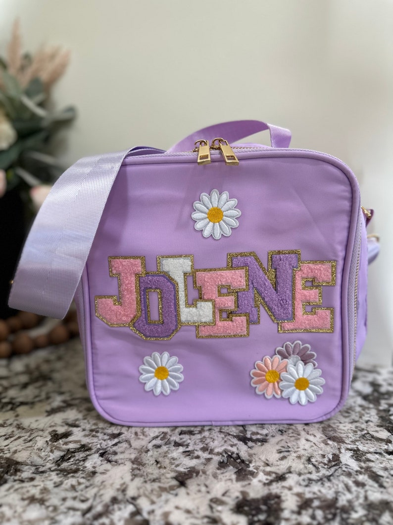 May include: A lavender lunch bag with a zipper closure and a personalized name "JOLENE" in pink, white, and gold letters. The bag is decorated with embroidered white daisies.