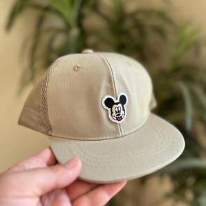 Könnte beinhalten: Eine beige und schwarze Baseballkappe mit einem Mickey Mouse-Aufnäher auf der Vorderseite.