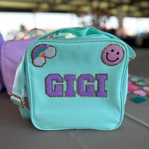 May include: A light blue lunch bag with a purple and gold "GIGI" name patch, a rainbow patch, and a pink smiley face patch.