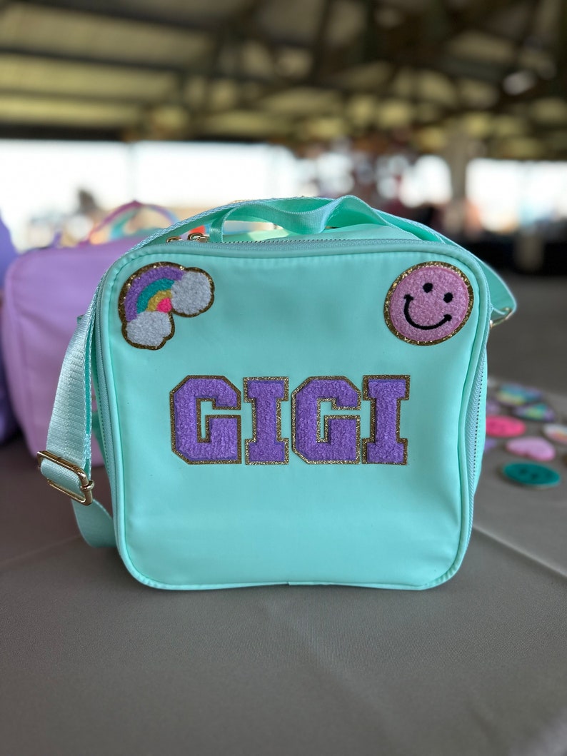 May include: A light blue lunch bag with a purple and gold "GIGI" name patch, a rainbow patch, and a pink smiley face patch.