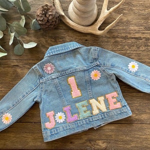 Könnte beinhalten: Eine hellblaue Jeansjacke mit einer pinkfarbenen, weißen und goldenen "1" und dem Namen "JOLENE" in bunten Buchstaben. Die Jacke ist mit weißen und rosa Blumen verziert.