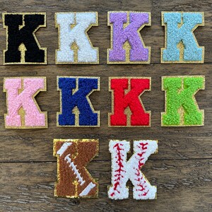 May include: Ten felt letter patches in various colors, including black, white, pink, blue, red, green, light blue, and purple. The letter is "K" in a varsity style font with a gold outline. Two additional patches feature a brown football and a white baseball with red stitching.