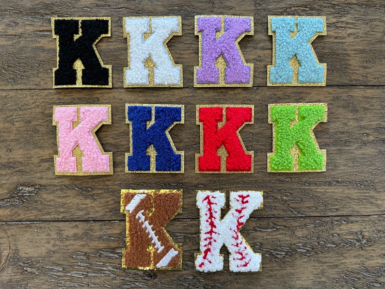 May include: Ten felt letter patches in various colors, including black, white, pink, blue, red, green, light blue, and purple. The letter is "K" in a varsity style font with a gold outline. Two additional patches feature a brown football and a white baseball with red stitching.