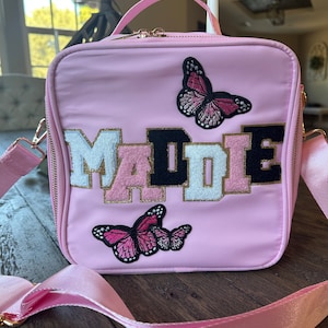 May include: A pink lunch bag with a personalized name "MADIE" in black, white, and gold letters. The bag has two butterfly appliques, one pink and one black.