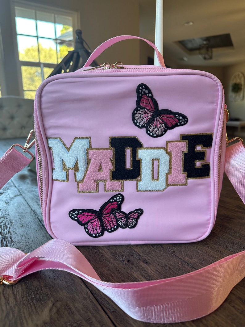 May include: A pink lunch bag with a personalized name "MADIE" in black, white, and gold letters. The bag has two butterfly appliques, one pink and one black.