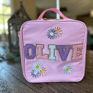 May include: Pink lunch bag with a gold zipper and the name "OLIVE" in colorful letters. The bag is decorated with embroidered flowers.
