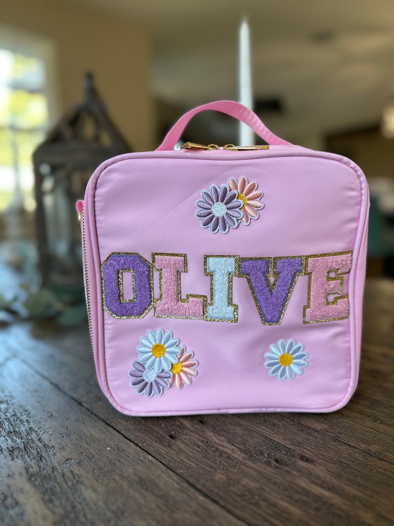 May include: Pink lunch bag with a gold zipper and the name "OLIVE" in colorful letters. The bag is decorated with embroidered flowers.