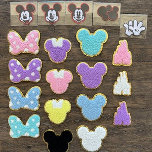 May include: A collection of 18 iron-on patches featuring Mickey Mouse, Minnie Mouse, and the Disney castle. The patches are in various colors, including pink, purple, blue, yellow, and white. Some patches are fuzzy and others are flat.