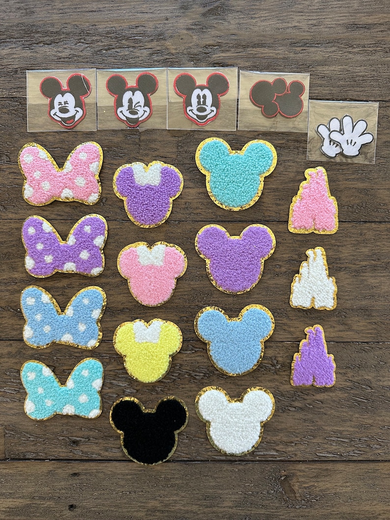 May include: A collection of 18 iron-on patches featuring Mickey Mouse, Minnie Mouse, and the Disney castle. The patches are in various colors, including pink, purple, blue, yellow, and white. Some patches are fuzzy and others are flat.
