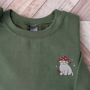 K&ouml;nnte beinhalten: Ein gr&uuml;ner Pullover mit einem gestickten Motiv eines wei&szlig;en Katers, der einen roten Pilz-Hut tr&auml;gt. Die Katze h&auml;lt einen kleinen Pilz in ihrer Pfote.