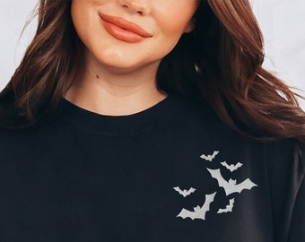 May include: Black crew neck sweatshirt with a small cluster of white bats on the upper left chest. The sweatshirt is paired with light blue jeans. A white, fluffy blanket is draped over the shoulders.
