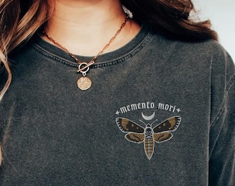 May include: Dark gray t-shirt with a moth and crescent moon design, and the words "Memento Mori". The shirt is paired with blue jeans and white sneakers. A gold necklace and bracelets are also visible.