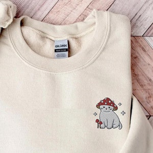 K&ouml;nnte beinhalten: Ein beiger Pullover mit einem kleinen gestickten Design eines Katers, der eine Pilzm&uuml;tze tr&auml;gt. Die Katze sitzt neben einem Pilz und es gibt kleine Sterne um sie herum.
