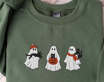 May include: An olive green crewneck sweatshirt featuring an embroidered design of three white ghosts. The first ghost wears an orange beanie and holds a black cat. The second ghost holds an orange pumpkin. The third ghost wears an orange scarf.