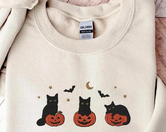 May include: A cream-colored crewneck sweatshirt featuring an embroidered design of three black cats sitting on orange jack-o'-lanterns, accompanied by small gold stars, a crescent moon, and two black bats, with a Gildan brand label at the neck.