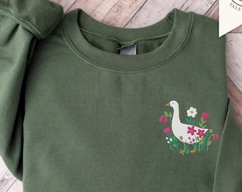 May include: An olive green crewneck sweatshirt featuring an embroidered design on the left chest. The embroidery depicts a white goose surrounded by small pink, red, and white flowers with green leaves. The sweatshirt has long sleeves with ribbed cuffs.