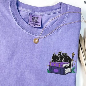 May include: A lavender Comfort Colors t-shirt featuring a small embroidered design of a black dragon sleeping on a stack of books. A gold necklace with a pendant is draped across the shirt. The shirt has a classic crew neck.