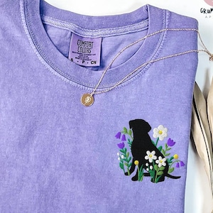 May include: A lavender short-sleeve t-shirt featuring an embroidered black silhouette of a dog surrounded by colorful flowers. The shirt has a crew neckline and a Comfort Colors label on the inside collar. A thin gold chain necklace rests on the fabric.
