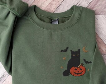 Op de afbeelding: Een groen sweatshirt met een zwarte kat die op een jack-o'-lantern zit. De kat heeft gele ogen en de jack-o'-lantern heeft een uitgesneden gezicht. Er zijn vleermuizen en sterren op de achtergrond.