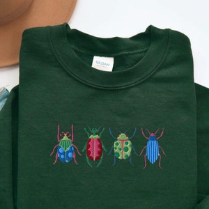 May include: A dark green sweatshirt with a row of four embroidered colorful beetles. The beetles are green, red, blue, and green with black accents.