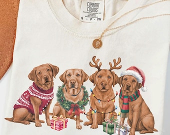 May include: A cream-colored t-shirt with a graphic of four Labrador Retrievers dressed in festive Christmas attire. The dogs are wearing a red sweater, a green wreath, Christmas lights, and a red and green scarf. The shirt has a tag that reads "Comfort Colors".