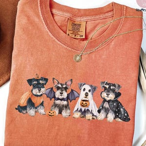 May include: An orange Comfort Colors t-shirt featuring four Schnauzer dogs dressed in Halloween costumes. The dogs are wearing a pumpkin costume, bat wings, a ghost costume, and a cape. A gold necklace with a pendant is also visible.