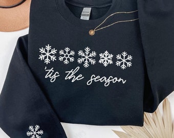 May include: Black sweatshirt featuring a white snowflake design and the phrase "'tis the season". A gold necklace with a pendant is draped on the sweatshirt. A single snowflake is on the sleeve. A tan hat is in the background.