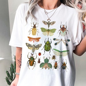 May include: White t-shirt with a colorful insect print. The insects include butterflies, beetles, and dragonflies. The shirt has a crew neck and short sleeves.