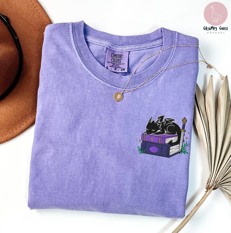 May include: A lavender Comfort Colors t-shirt featuring a small embroidered design of a black dragon sleeping on a stack of books. A gold necklace with a pendant is draped across the shirt. The shirt has a classic crew neck.
