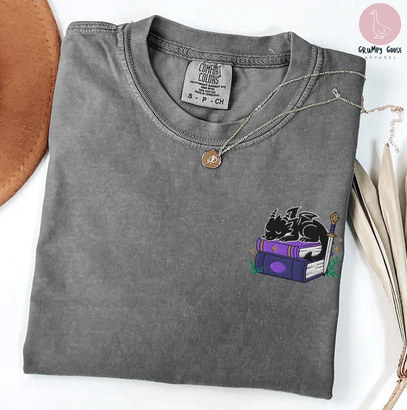 May include: A gray Comfort Colors t-shirt with a small embroidered design of a black dragon sleeping on purple books. A silver necklace with a pendant rests on the shirt. The shirt is size S-P-CH.