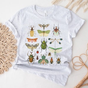 May include: A light gray t-shirt with a vintage insect illustration print. The print features a variety of insects, including butterflies, moths, beetles, and dragonflies. The insects are depicted in a variety of colors, including green, brown, red, and yellow.