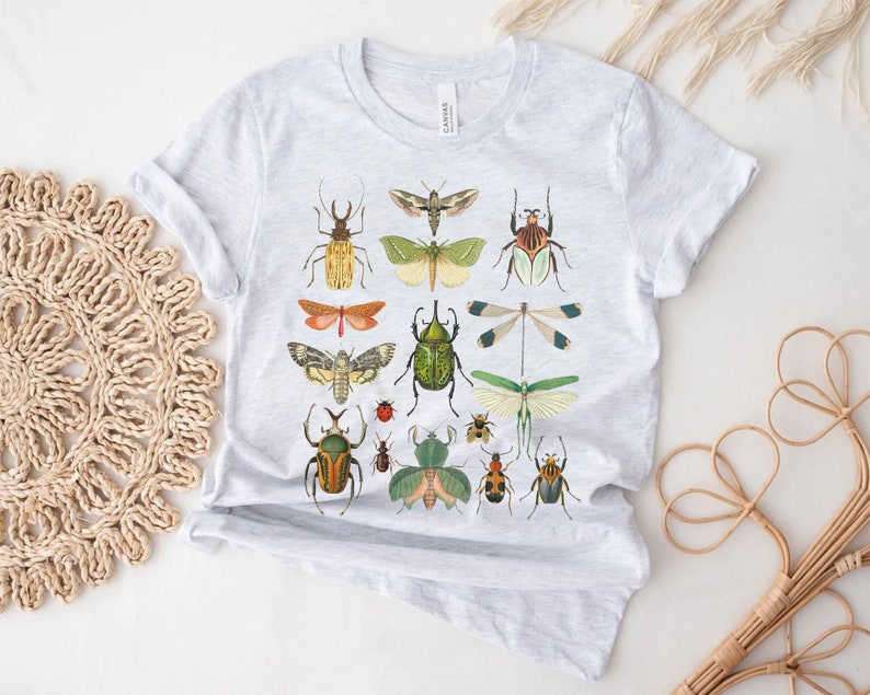 May include: A light gray t-shirt with a vintage insect illustration print. The print features a variety of insects, including butterflies, moths, beetles, and dragonflies. The insects are depicted in a variety of colors, including green, brown, red, and yellow.